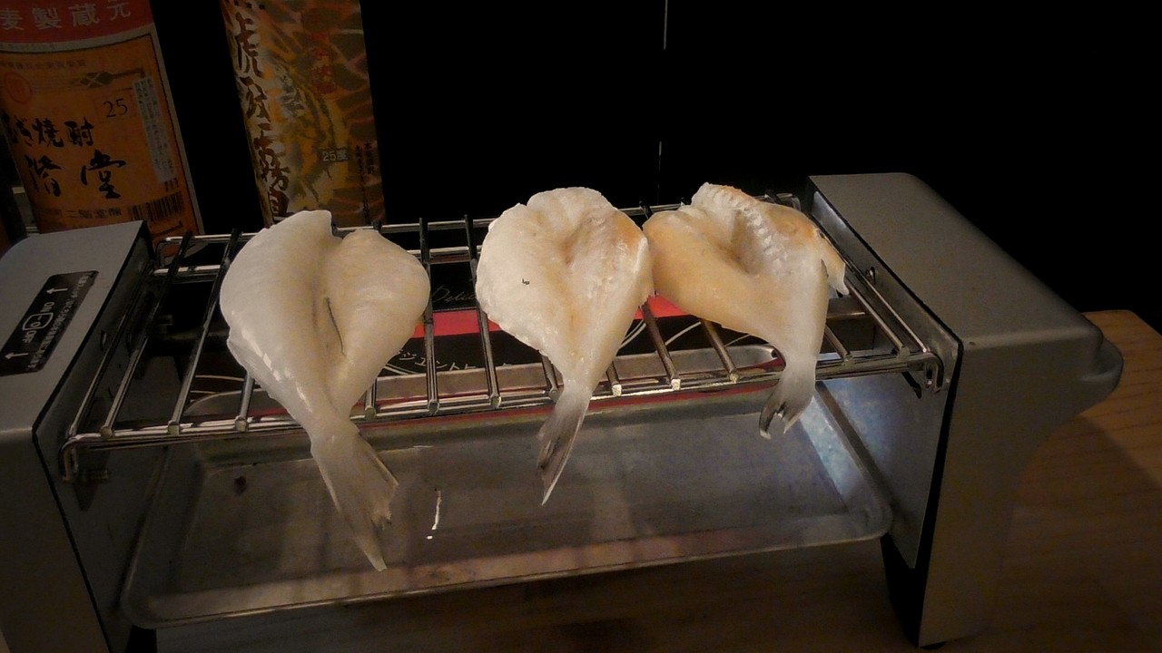 焼き鳥用の電気グリルの網に、ふぐの一夜干しが3枚ならんでいる。奥に焼酎のビン