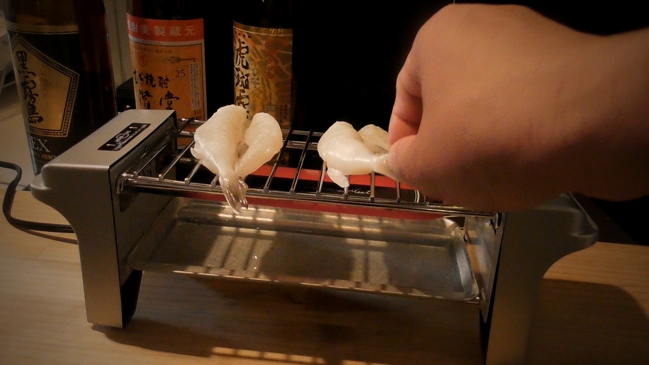 焼き鳥グリルの網に白いふぐの一夜干しを1枚のせ、右手で2枚目をのせているところ。奥に焼酎のビン