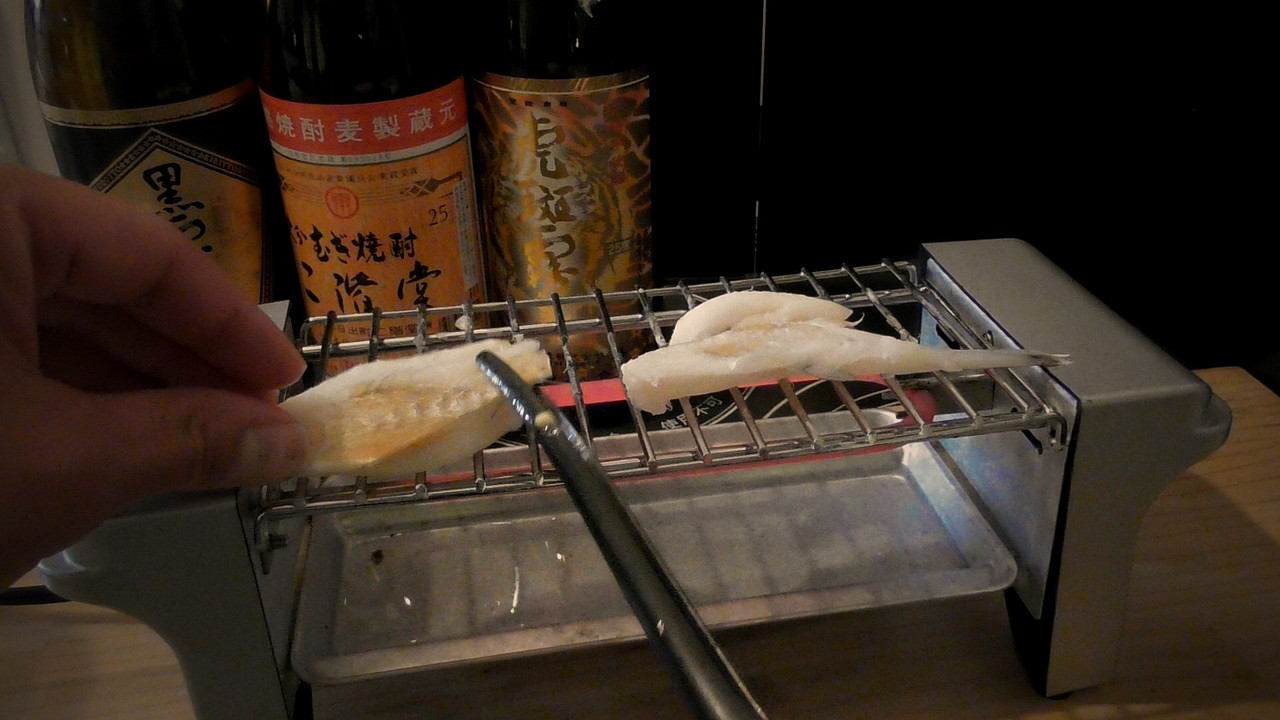 網の上の焼けたふぐを左手で押さえ、黒いトングで身をほぐしているところ。右にもう1枚。奥に焼酎のビン