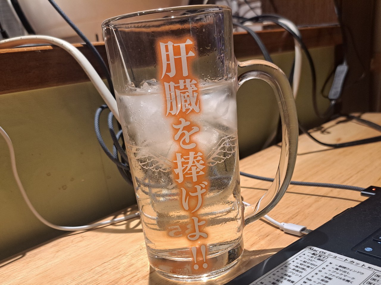 机の上の、取っ手つきのガラスの中ジョッキ。胴にオレンジ色の縦書きで「肝臓を捧げよ!!」、左右に白い線の翼の紋章。中は氷と透明な飲み物で、表面に水滴。後ろに木の棚とコード、右下にノートパソコンのキーボードの角
