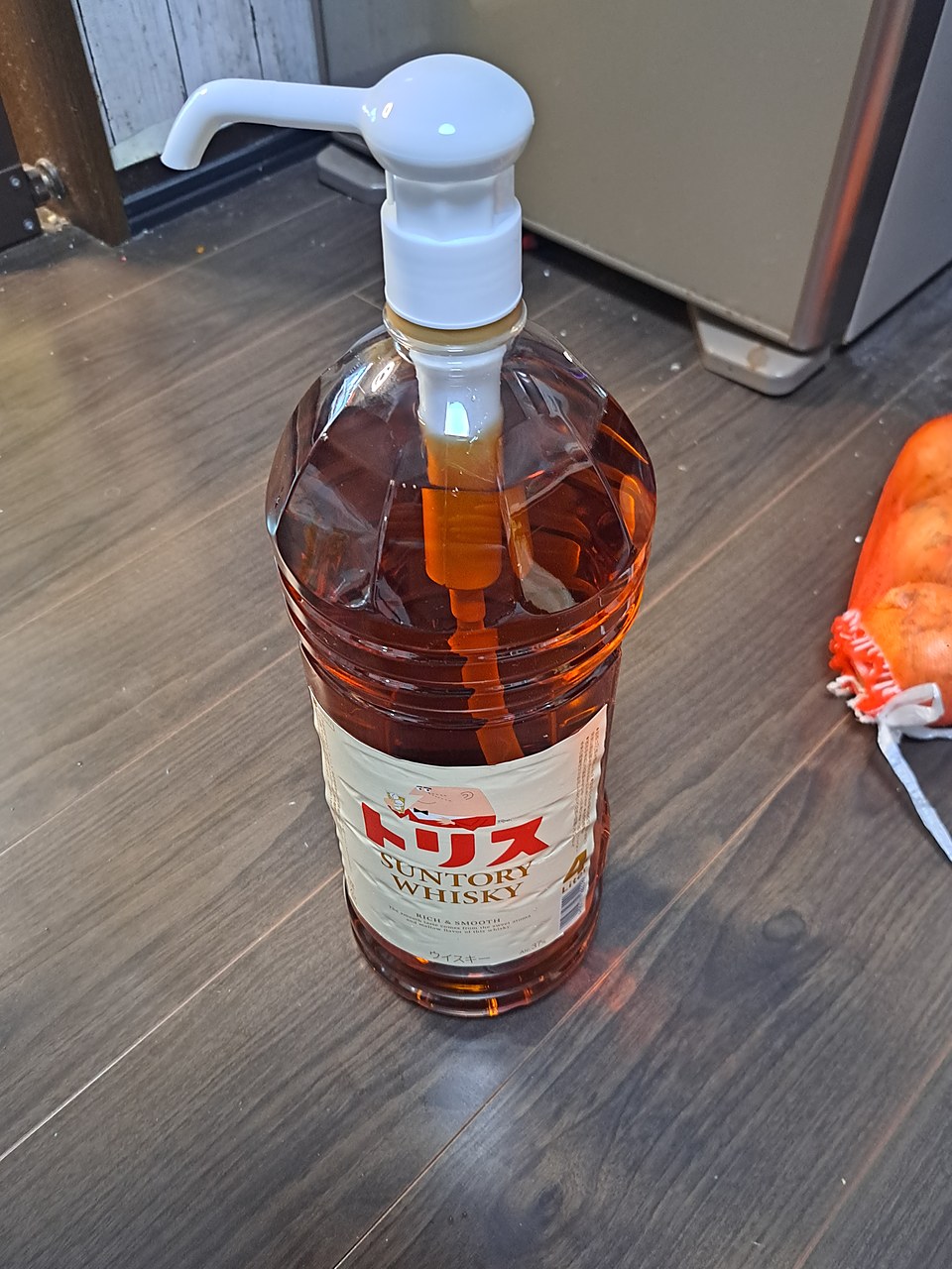木目の床に立つ、サントリー「トリス」の4Lペットボトル。キャップのかわりに白いポンプが付いていて、ボトルの中にポンプから下りたチューブが透けて見える。ラベルにはグラスを持ったおじさんの絵と大きな「4」。右はしにオレンジ色のネット袋