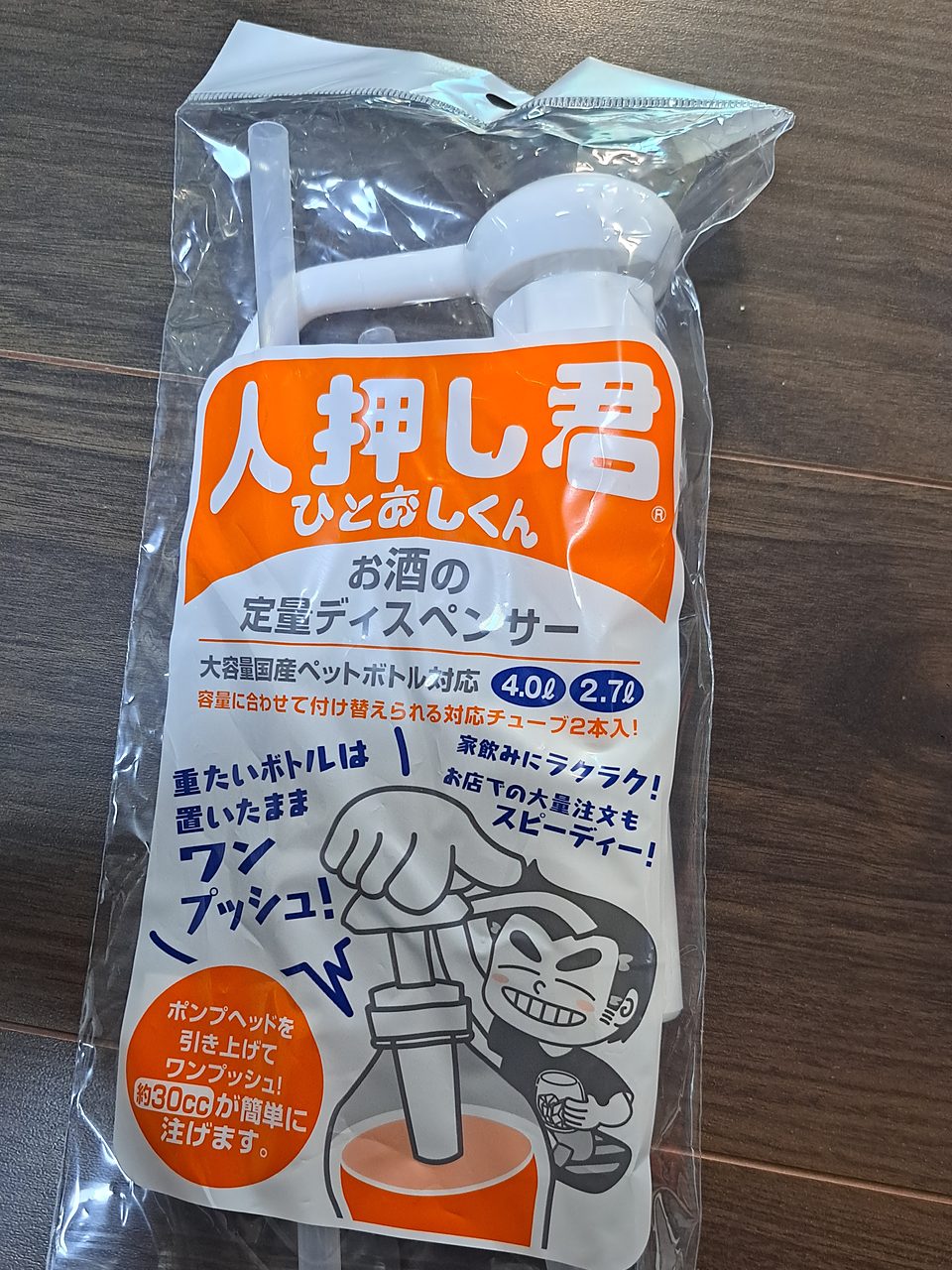 床に置いたポンプの袋の表。オレンジ色の大きな文字で「人押し君」、ふりがな「ひとおしくん」と®マーク。その下に「お酒の定量ディスペンサー」「大容量国産ペットボトル対応 4.0ℓ 2.7ℓ」。大きな手がポンプを押すイラストと、グラスを持って笑う男の人のイラスト