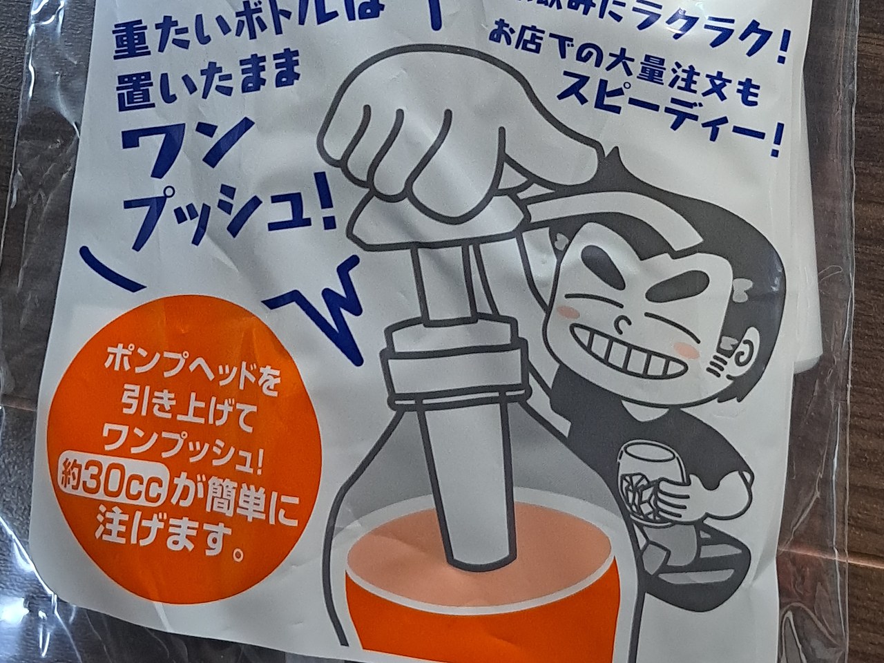 袋の表のアップ。オレンジ色の丸の中に白い文字で「ポンプヘッドを引き上げてワンプッシュ！約30ccが簡単に注げます。」。上に「重たいボトルは置いたまま ワンプッシュ！」、右にグラスを持って笑う男の人のイラスト