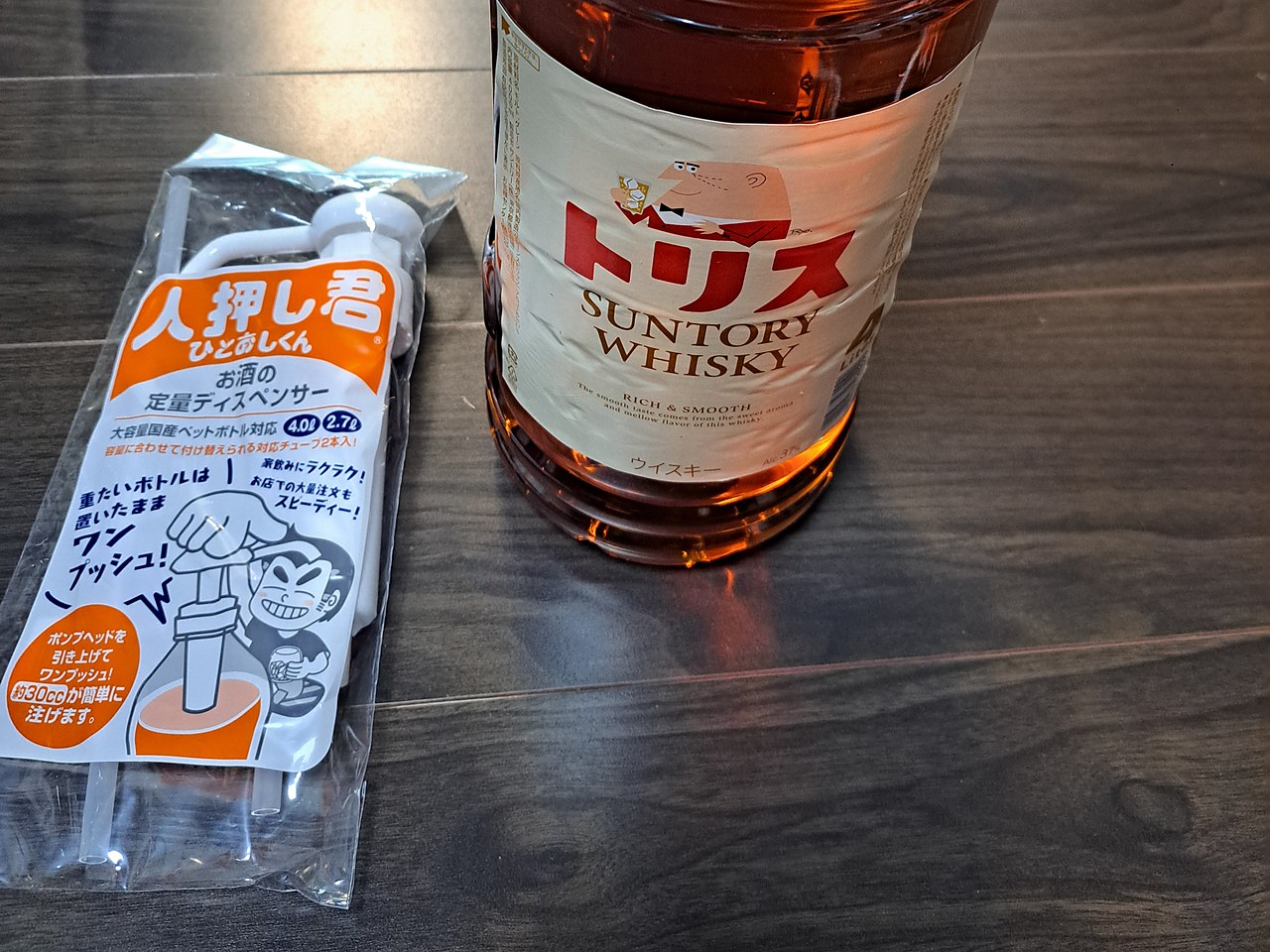 木目の床に、ポンプの入った袋と、トリスの4Lペットボトルが並んでいる。袋にはオレンジ色の「人押し君」の文字とイラスト、ボトルのラベルには「トリス SUNTORY WHISKY」とグラスを持ったおじさんの絵